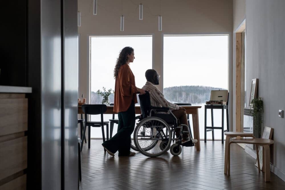 A woman pushes a man in a wheelchair through a dining area with large windows and a view of the outdoors. - Home Instead Southampton