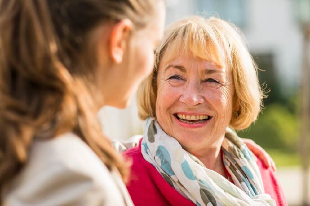 Two women outdoors smiling at each other, one older woman with short blonde hair and a scarf, the other younger with long hair. - Home Instead Southampton