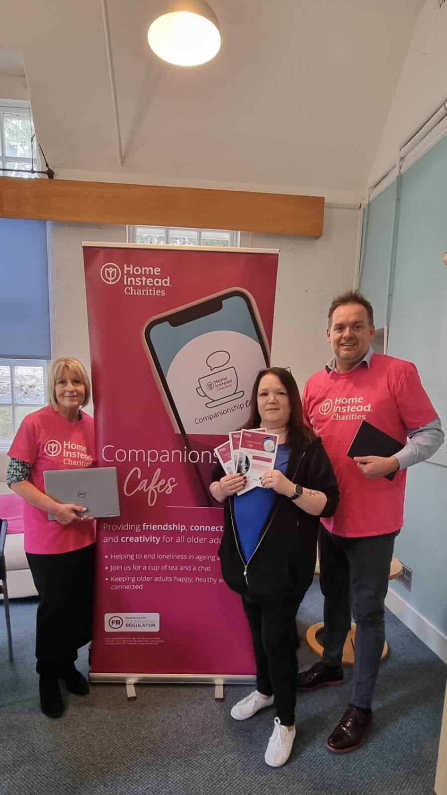 Three people in pink shirts stand by a "Home Instead Charities" banner, holding promotional materials. - Home Instead