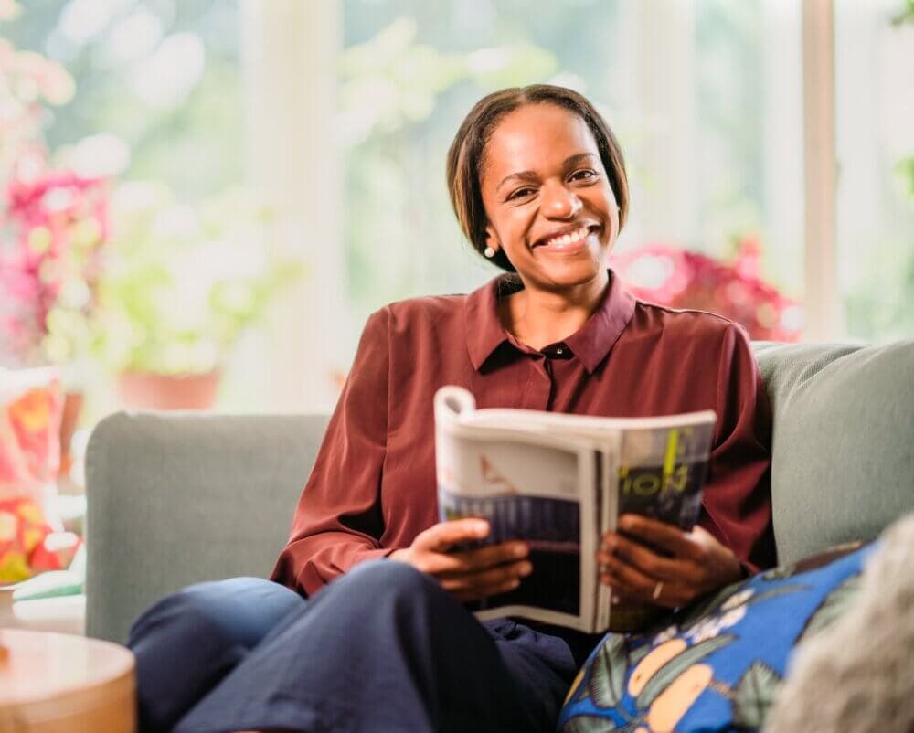 A smiling woman in a maroon blouse sits on a couch, holding an open book, with bright sunlight in the background. - Home Instead