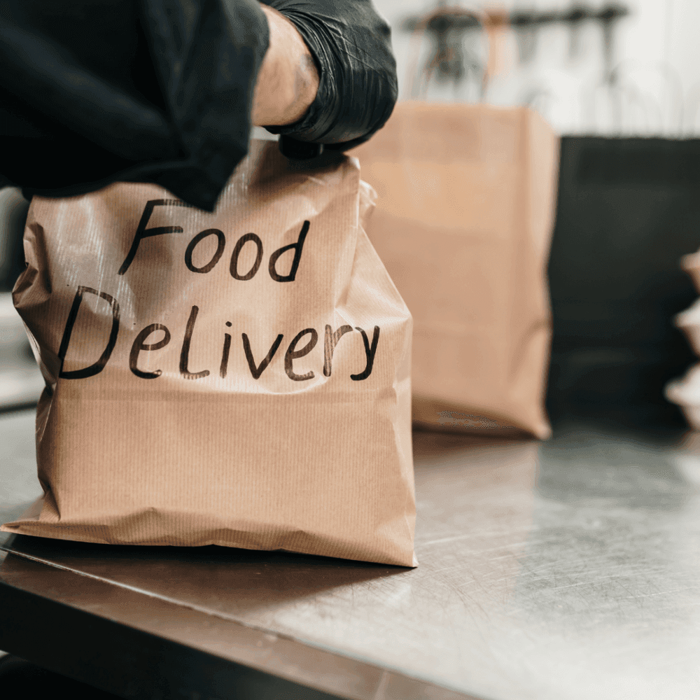 Person wearing gloves holding a brown paper bag labeled "Food Delivery" in a kitchen setting. - Home Instead