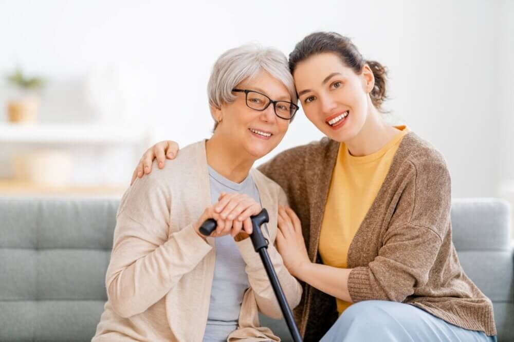 A senior woman and a younger woman, both smiling, sit on a couch together, sharing a warm embrace. - Home Instead