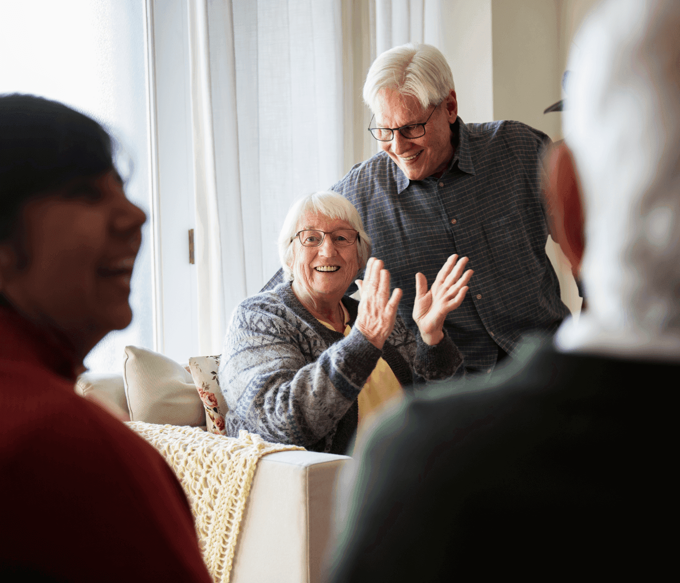 A group of elderly people enjoy a lively conversation in a warmly lit room, one woman claps and smiles. - Home Instead