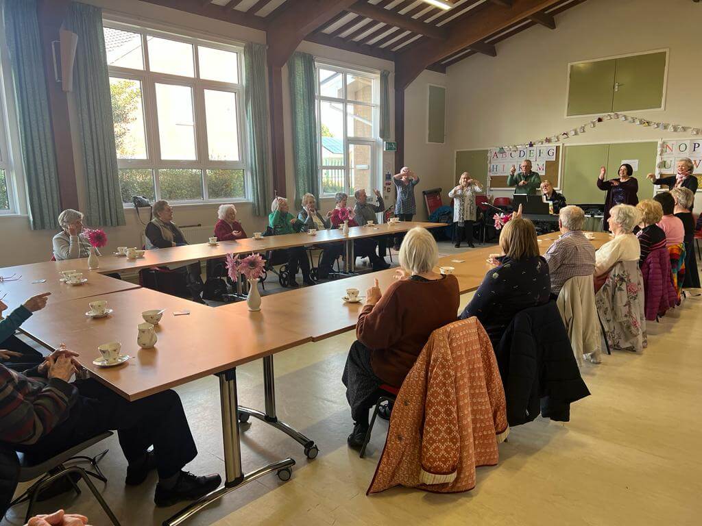 A group of elderly people sit at tables in a community hall, engaging in a discussion or activity. - Home Instead