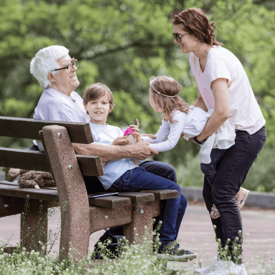 Four people enjoying a day in the park, with two children and two adults, one elderly, sitting and interacting happily. - Home Instead