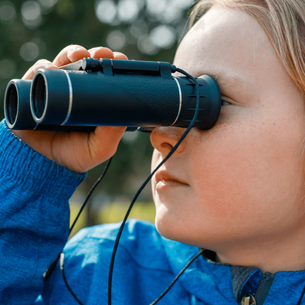 A child wearing a blue jacket looks through binoculars with a focused expression. - Home Instead