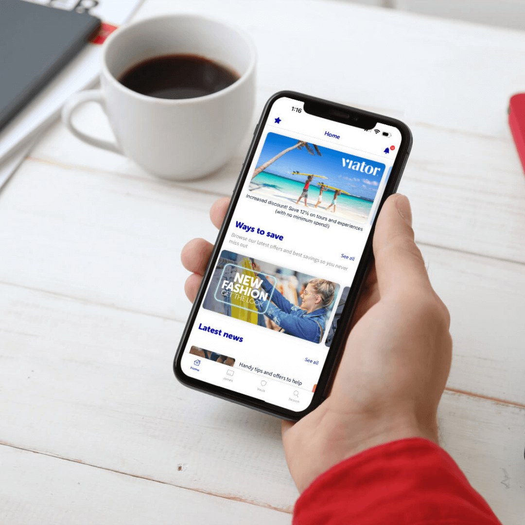 Person holding a smartphone displaying a travel app while having a cup of coffee on a desk with a notebook. - Home Instead