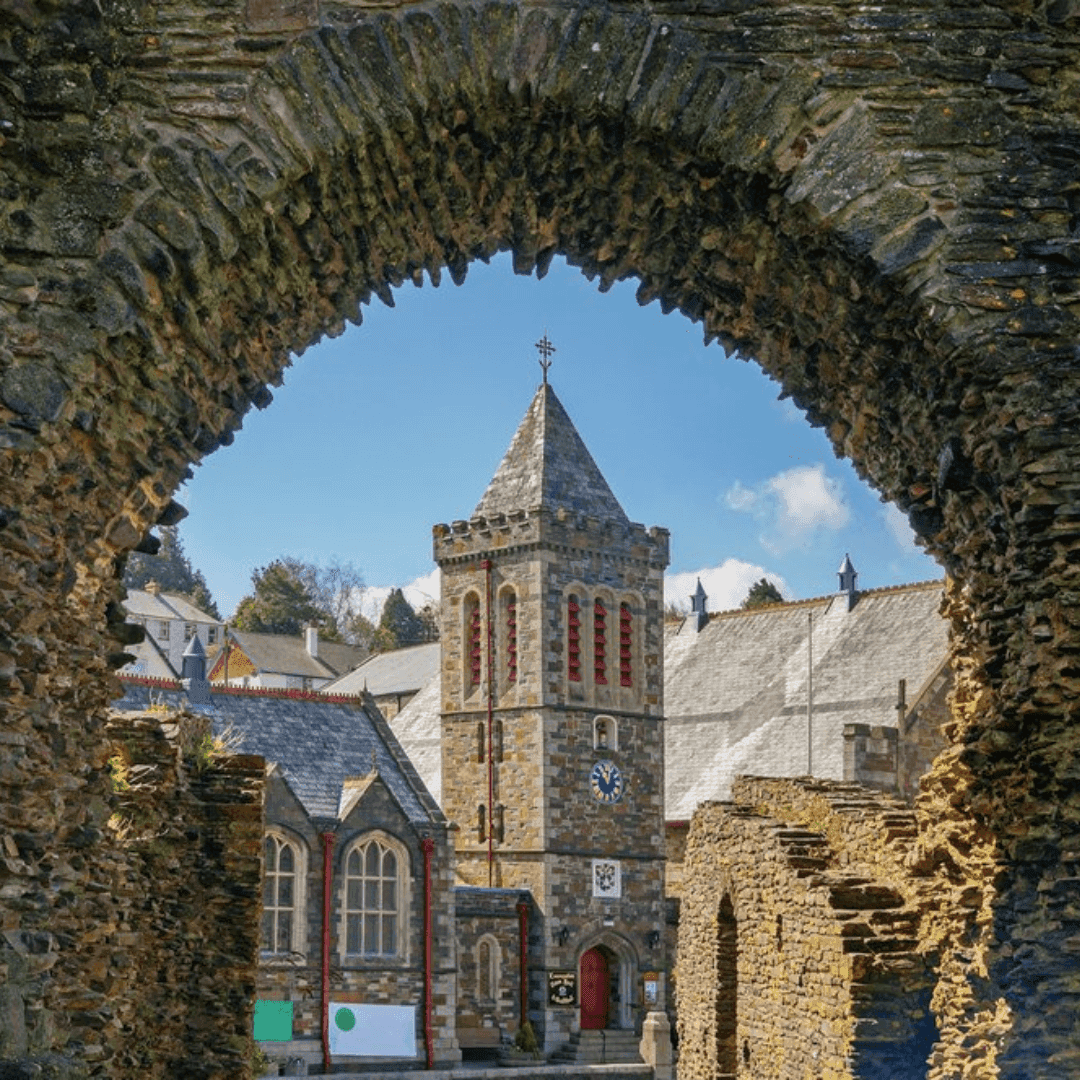 A stone church tower seen through a stone arch, surrounded by historic buildings under a blue sky. - Home Instead