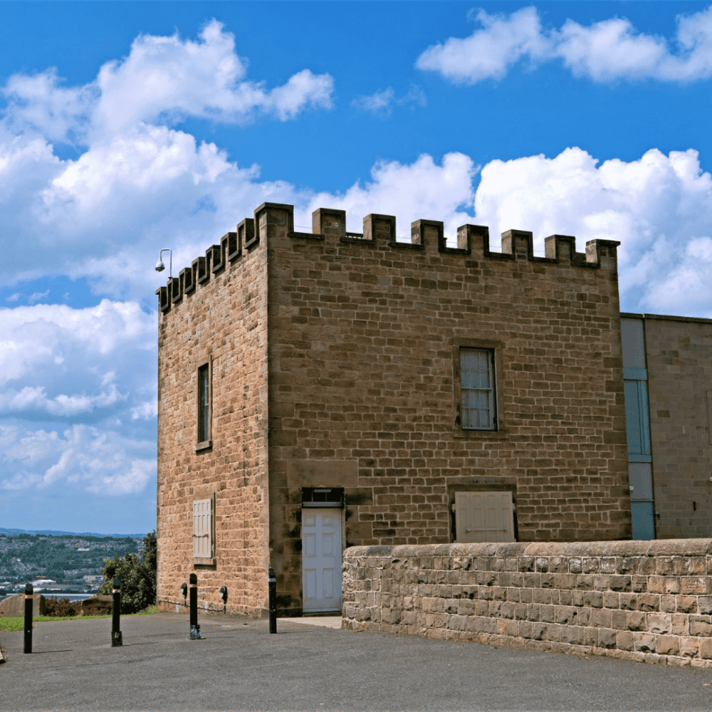 A historic stone building with crenellations, under a bright blue sky with clouds, set against a scenic landscape. - Home Instead