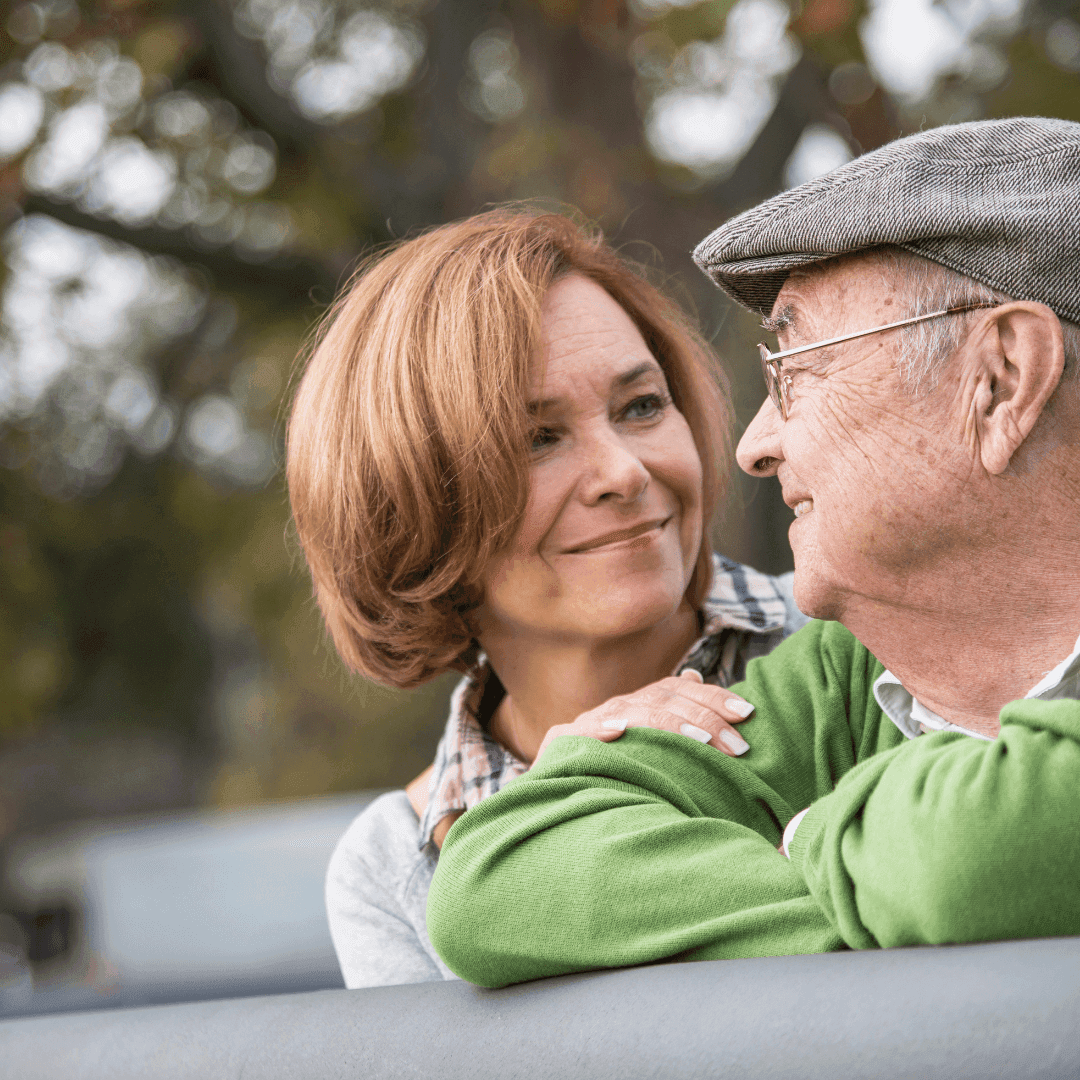 An elderly man and a middle-aged woman share a warm moment outdoors, smiling at each other. - Home Instead