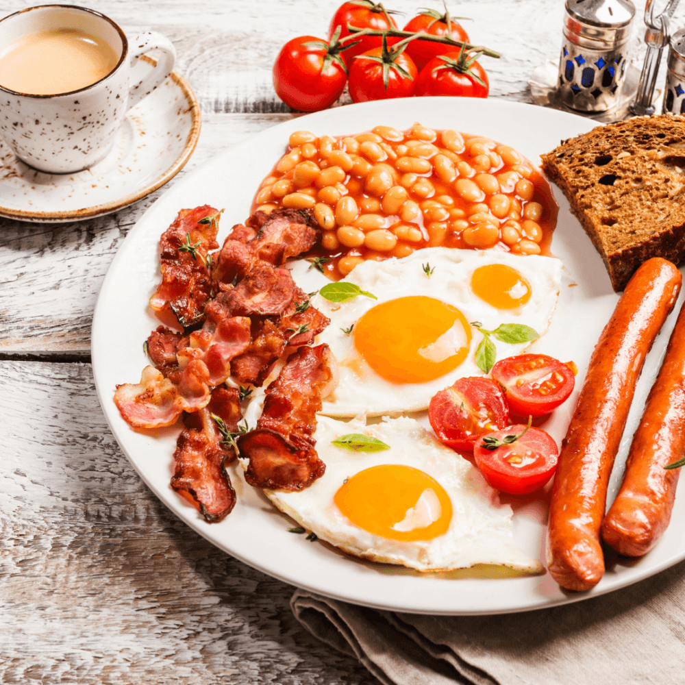 A plate with fried eggs, bacon, sausages, baked beans, tomatoes, and toast, next to a cup of coffee on a rustic table. - Home Instead