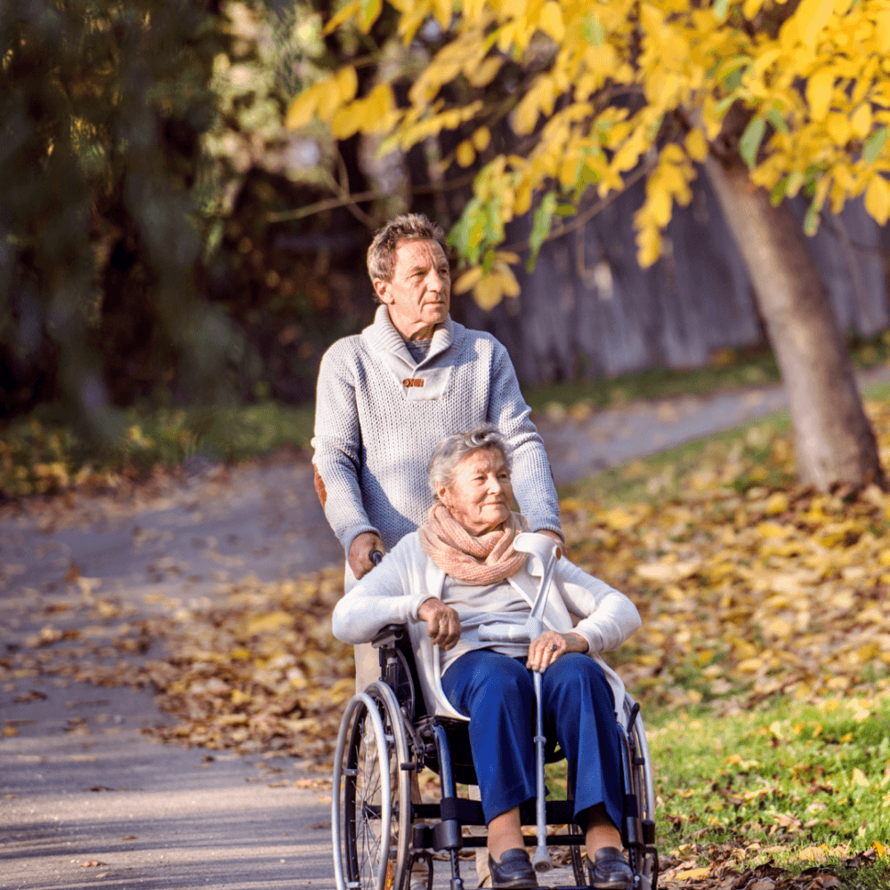 A man pushes a woman in a wheelchair along a tree-lined path with fallen autumn leaves. - Home Instead