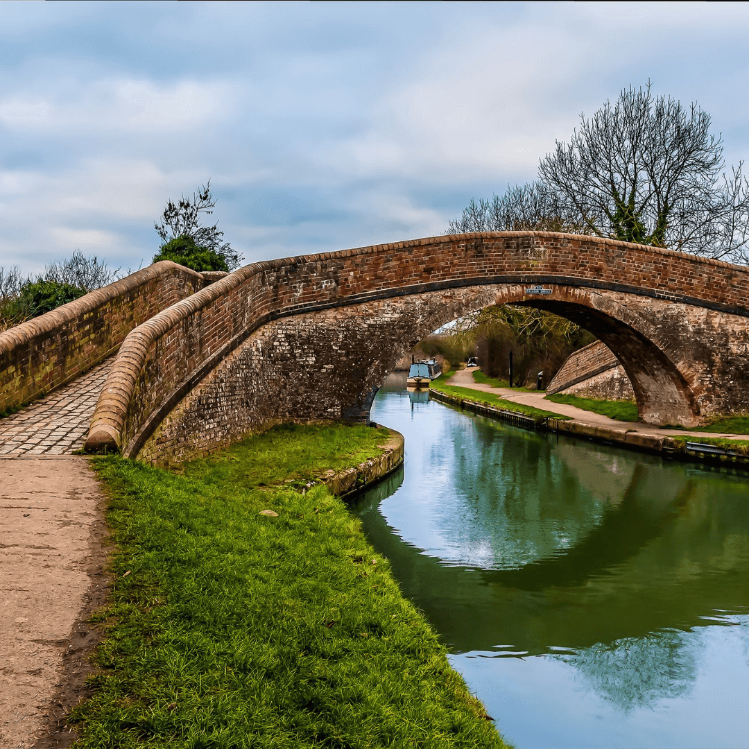 Arched brick bridge over a serene canal, with a path on the left side and green grass along the canal's edge. - Home Instead