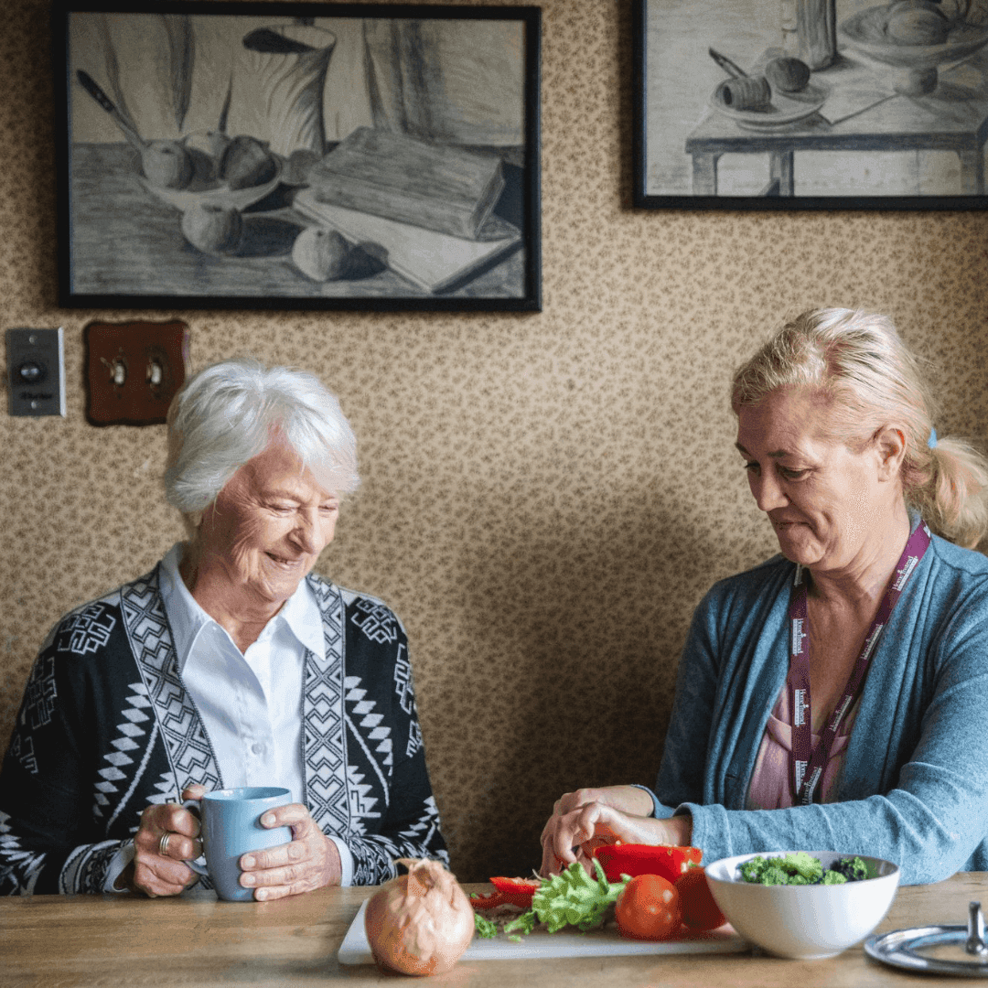 Two women at a kitchen table: one drinks from a mug, while the other cuts vegetables. Artwork hangs on the wall behind them. - Home Instead