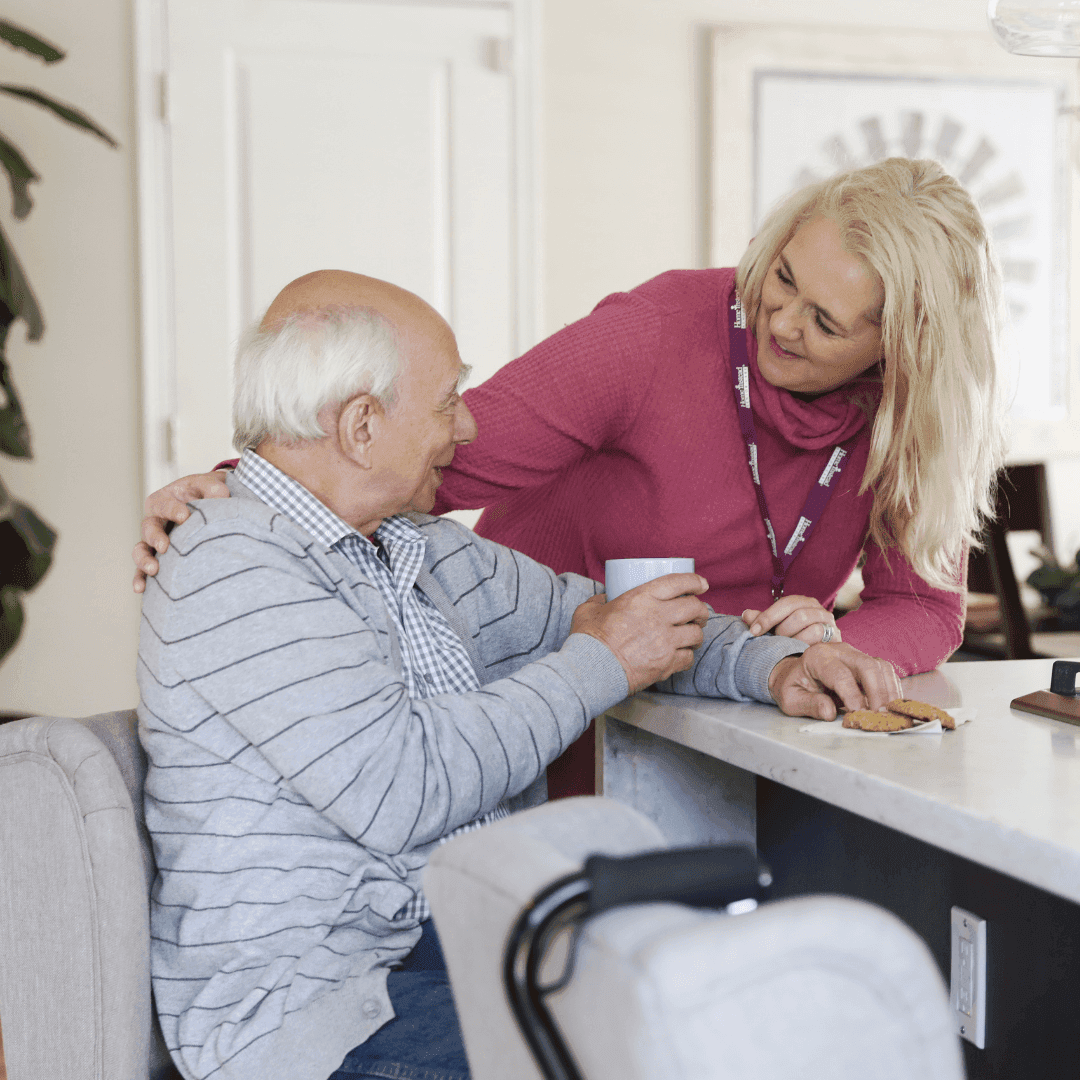 A caregiver in a pink sweater helps an elderly man with a snack and drink at a kitchen counter. - Home Instead