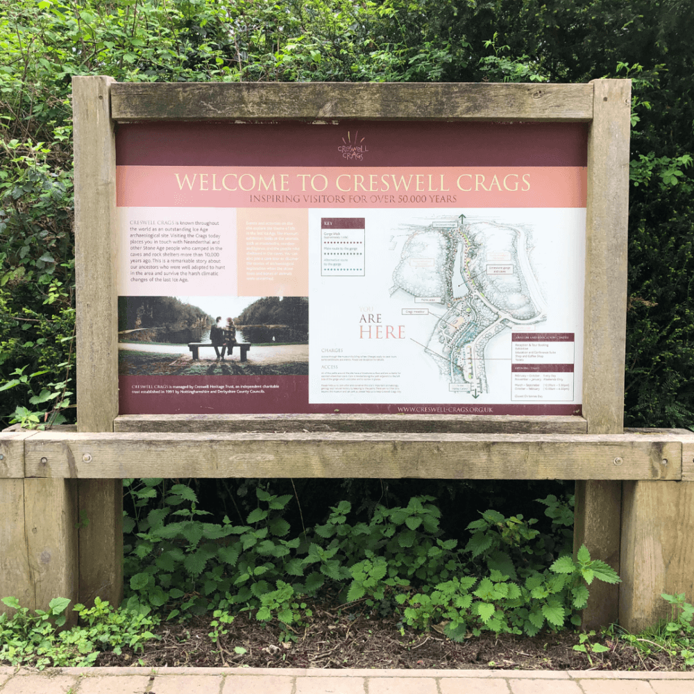 Signboard surrounded by greenery, titled "Welcome to Creswell Crags" with a map and information for visitors. - Home Instead