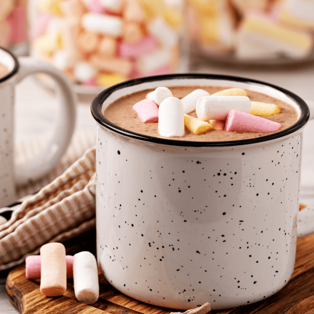 A white speckled mug of hot chocolate topped with colorful mini marshmallows, with another mug and marshmallows in the background. - Home Instead