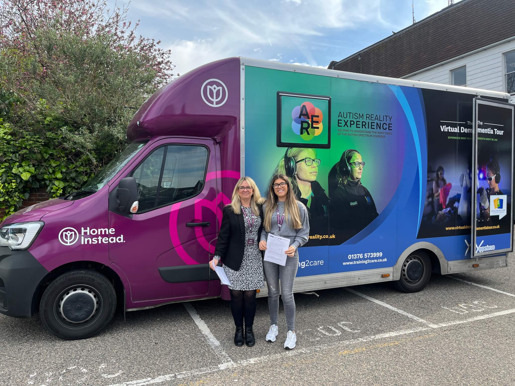 Two people standing in front of a colorful van with "Autism Reality Experience" and "Home Instead" branding. - Home Instead