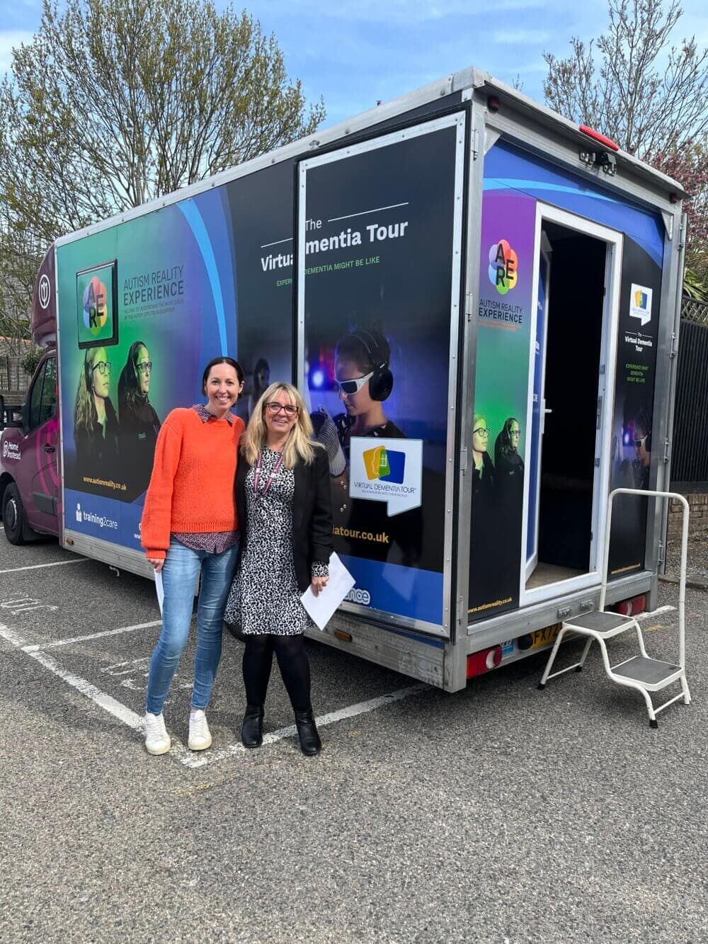 Two people smiling and standing in front of a colorful virtual dementia tour truck in a parking lot. - Home Instead