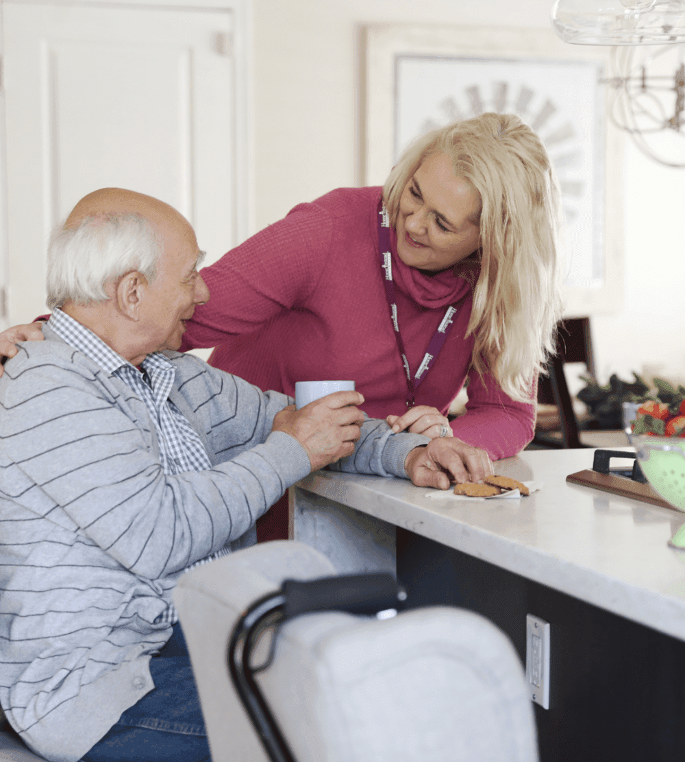 Elderly man and woman sharing a warm moment at a kitchen island, man holding a mug, woman smiling and leaning on his shoulder. - Home Instead