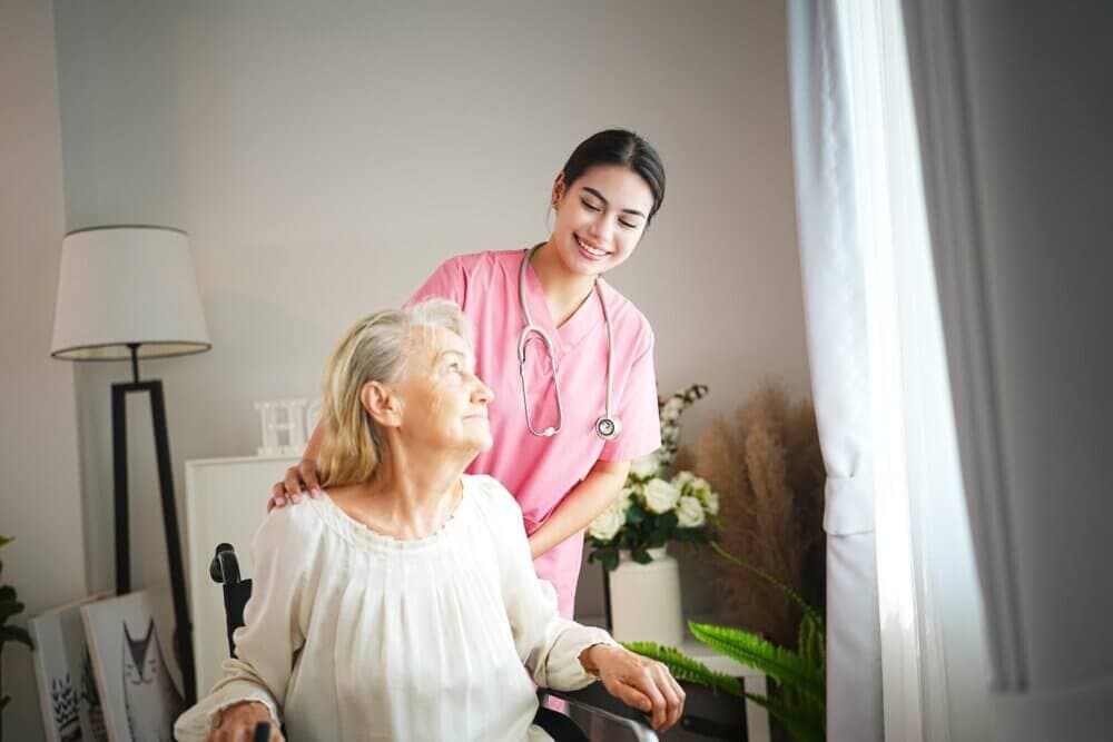 A nurse in pink scrubs smiles while assisting an elderly woman seated in a wheelchair near a window. - Home Instead