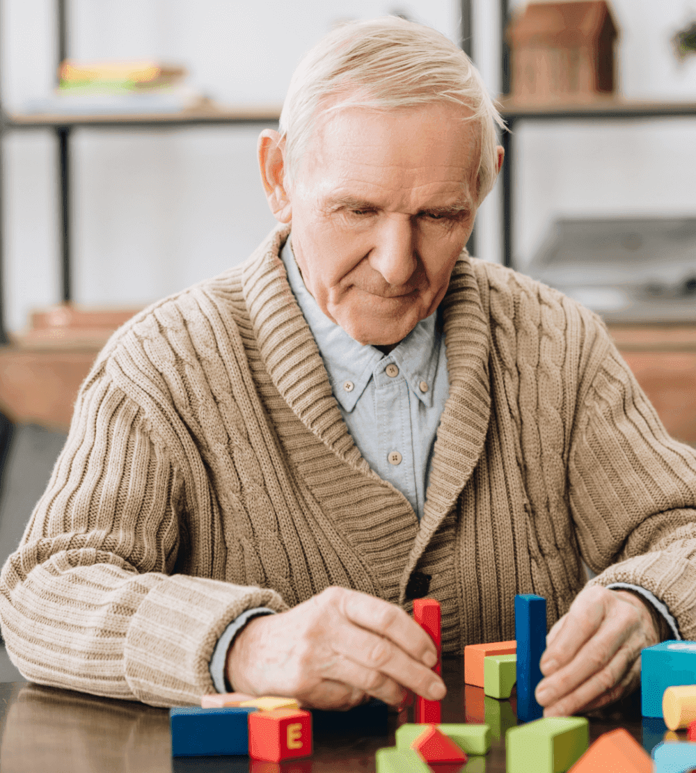An elderly man wearing a tan sweater plays with colorful building blocks on a table. - Home Instead