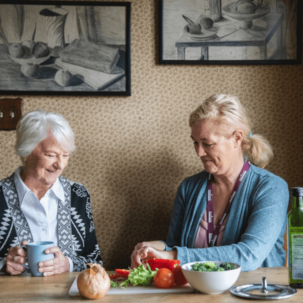 An elderly woman and a caregiver are preparing vegetables together in a cozy kitchen. - Home Instead