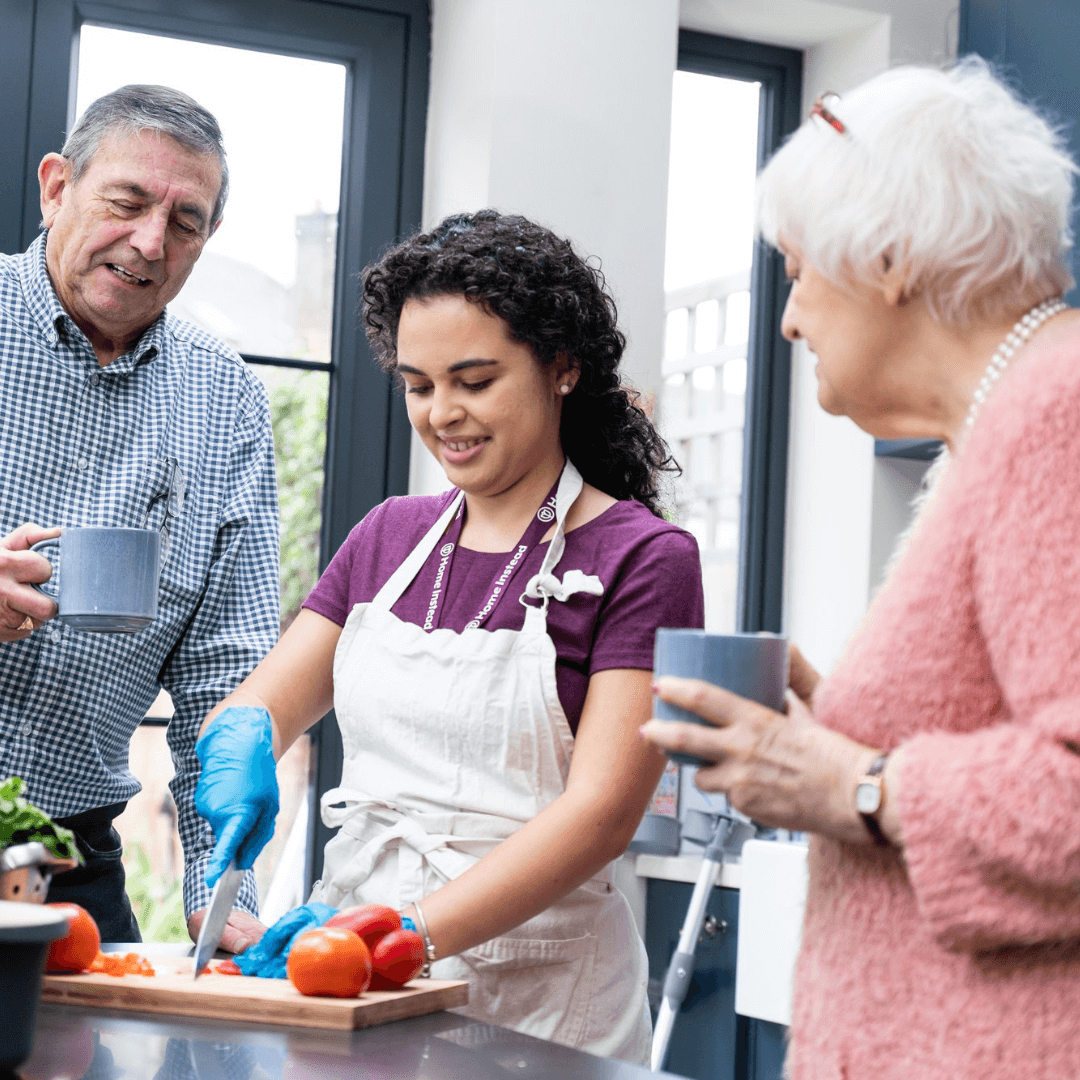 Young woman chopping vegetables while two older individuals watch and drink from mugs in a bright kitchen. - Home Instead