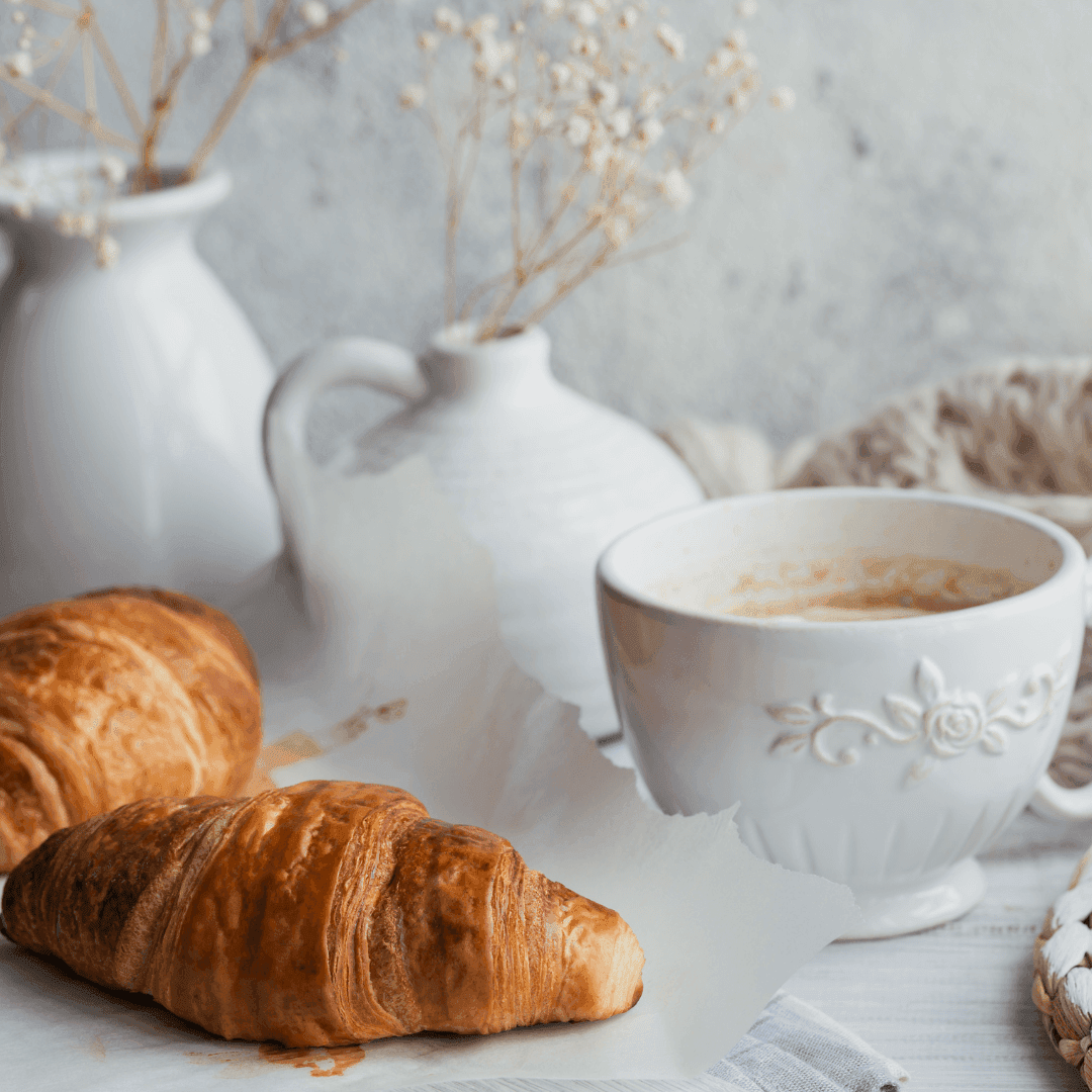 Two croissants on parchment paper with a cup of coffee beside white ceramic vases holding dried flowers in the background. - Home Instead