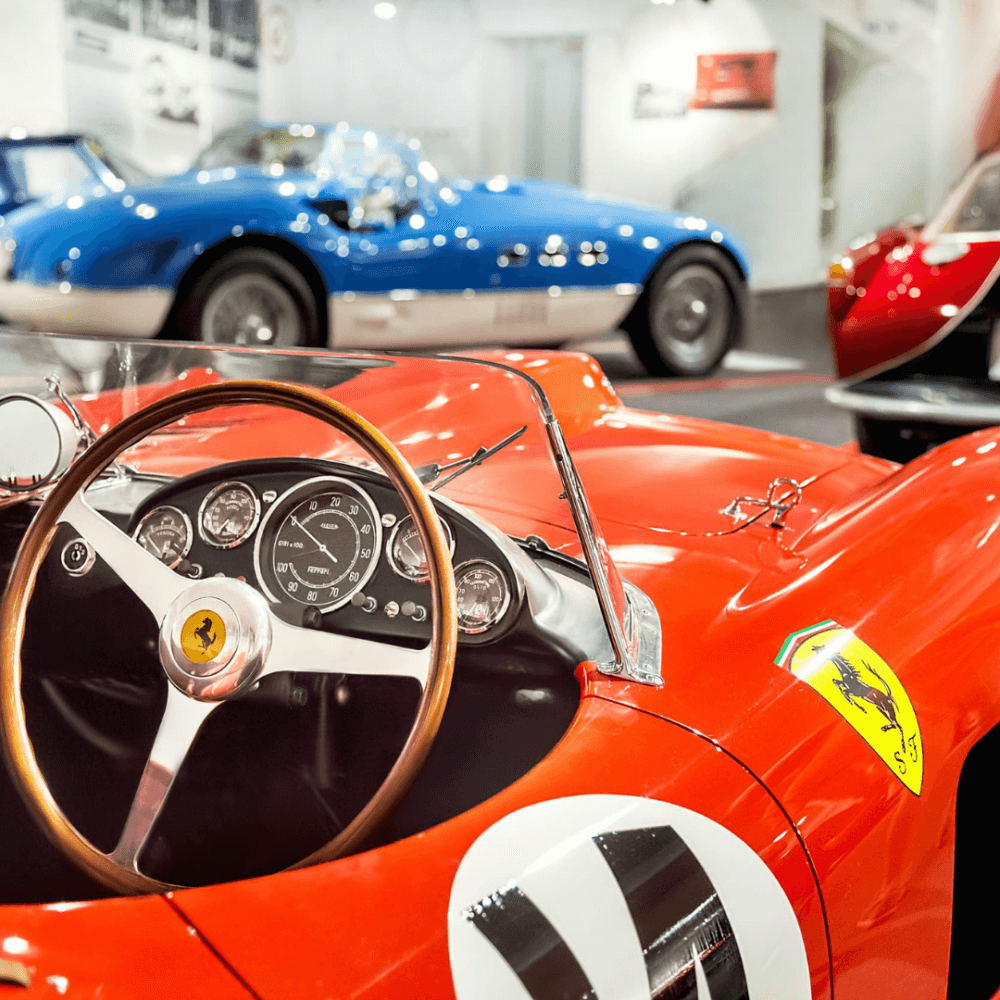 Close-up of a vintage red Ferrari dashboard and steering wheel, with blue cars visible in the background. - Home Instead