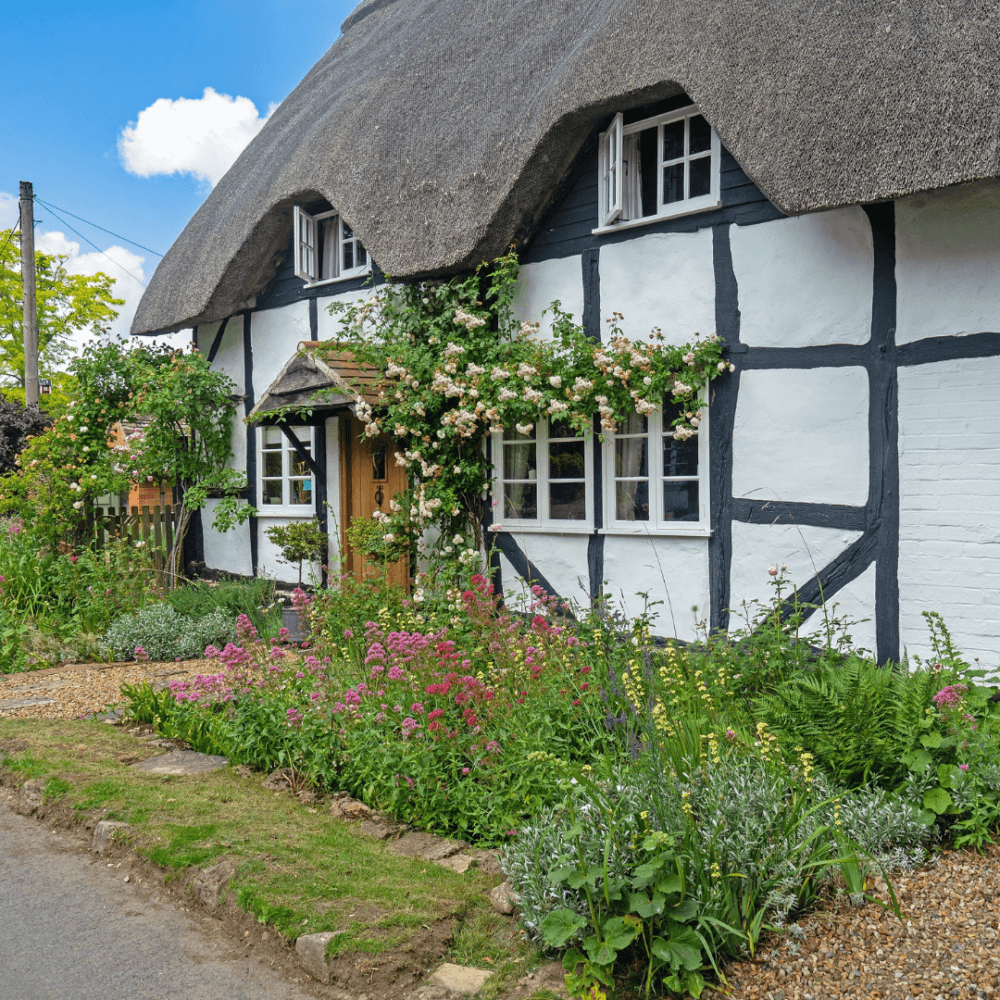 Quaint cottage with a thatched roof, white walls, black timber framing, and a lush flower garden along a narrow path. - Home Instead