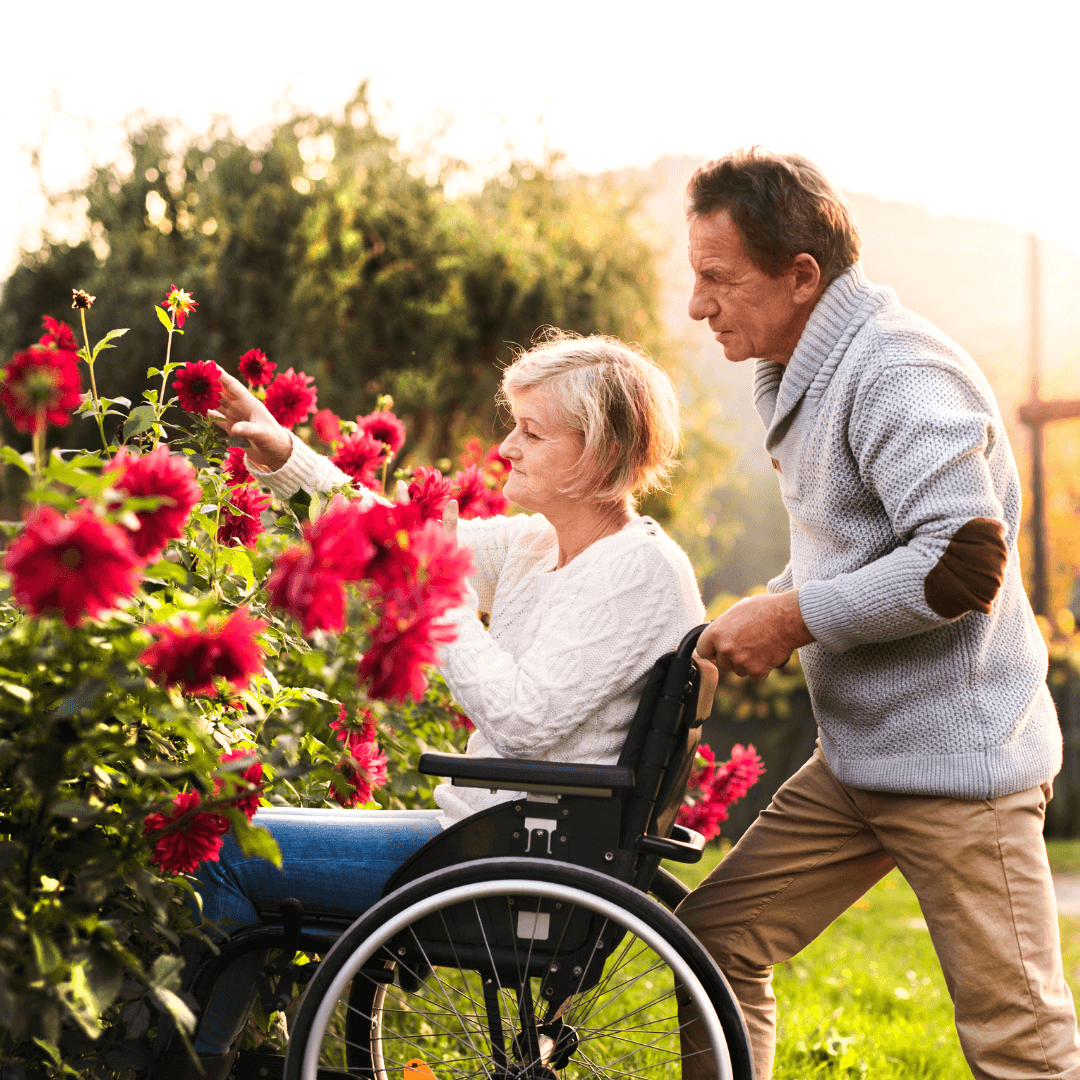 A woman in a wheelchair reaches for flowers in a garden while a man stands behind her, assisting. - Home Instead