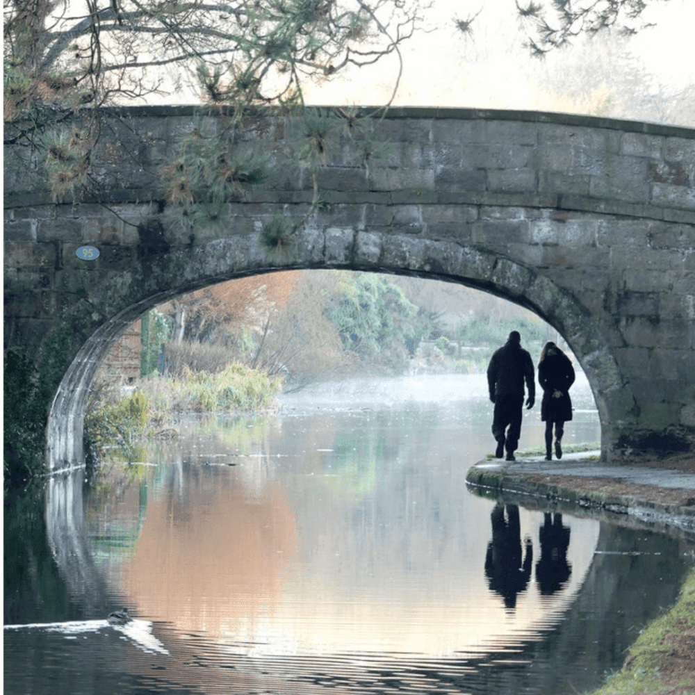 Two people walk under a stone bridge beside a calm river reflecting the surrounding trees and sky. - Home Instead