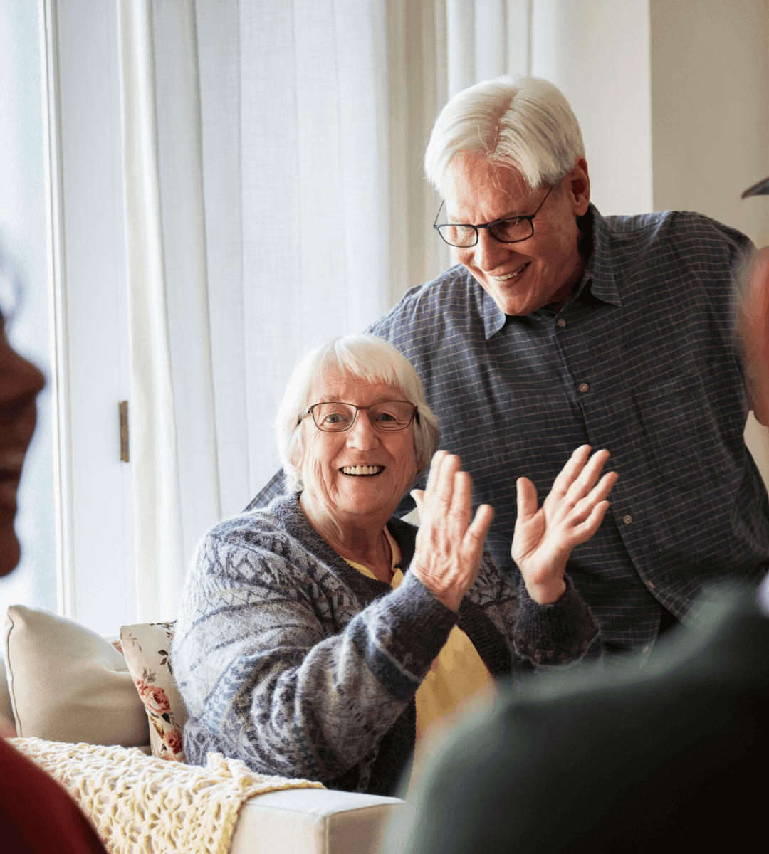 An elderly woman sitting and clapping, next to a smiling elderly man wearing glasses, in a well-lit room. - Home Instead