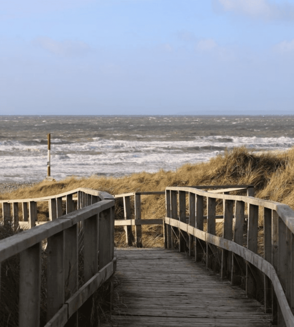 Wooden boardwalk leading through dunes to a beach with rough waves and a cloudy sky in the background. - Home Instead