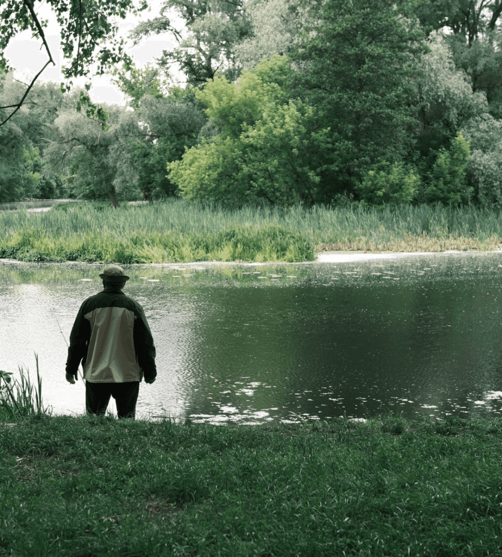 Person in a jacket and hat stands by a calm river, surrounded by lush green trees and foliage. - Home Instead