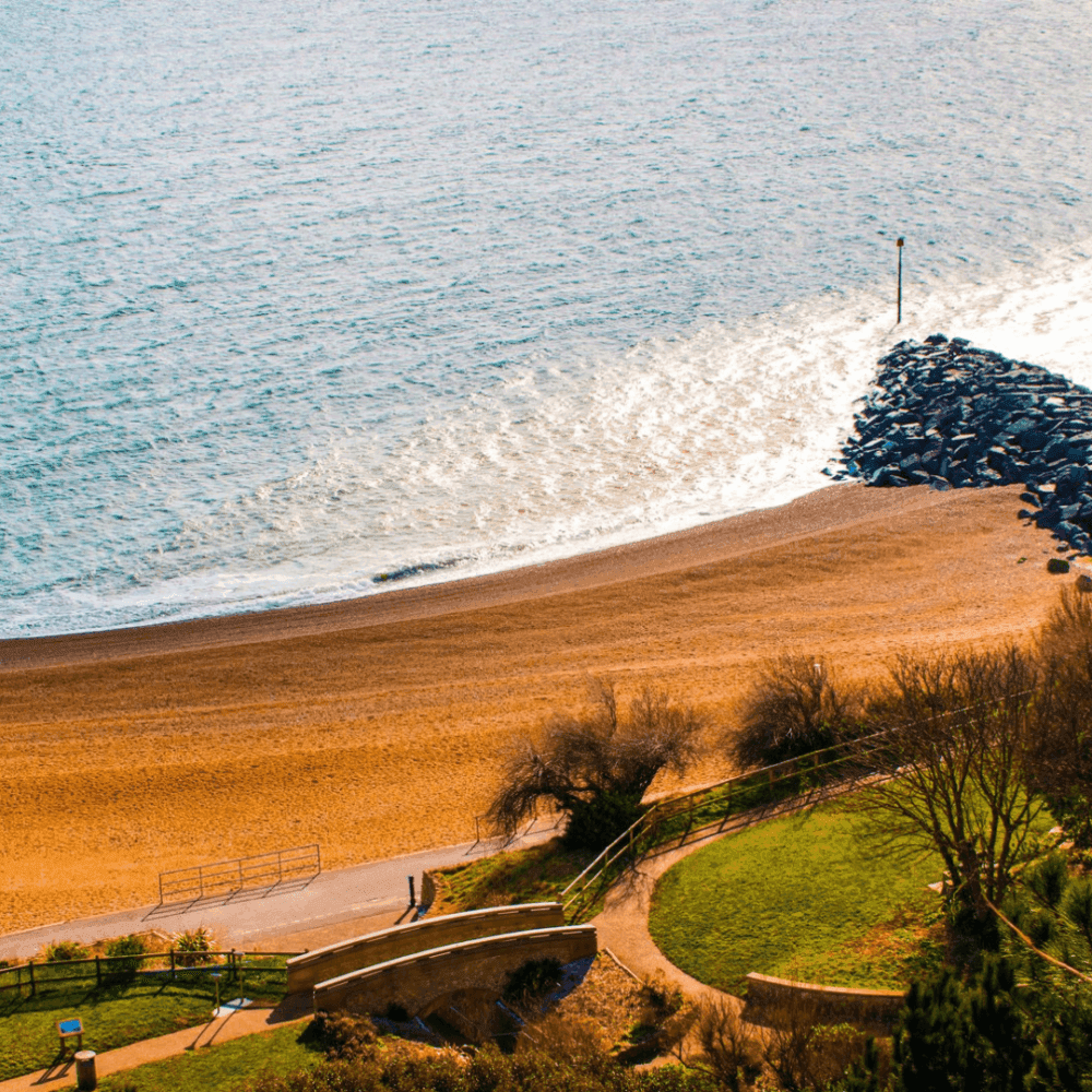 Elevated view of a serene beach with golden sand, gentle waves, a breakwater, and a grassy area with a walkway. - Home Instead