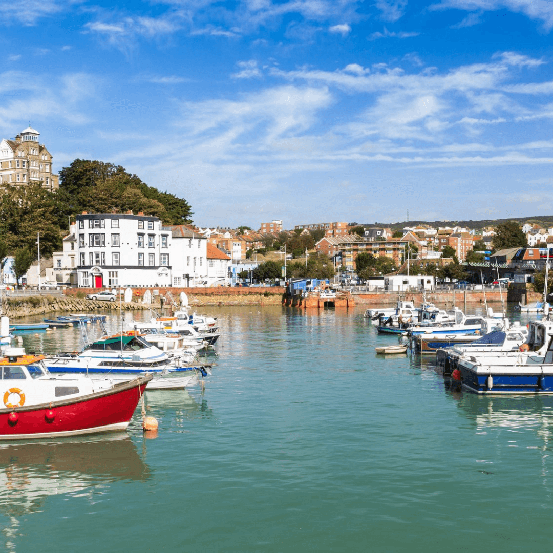 Boats docked in a calm harbor with buildings and trees in the background under a bright blue sky with scattered clouds. - Home Instead
