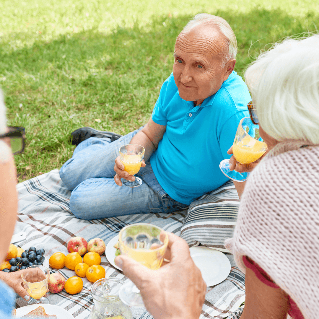 Three elderly people enjoying a picnic on a blanket with fruits and drinks in a park. - Home Instead