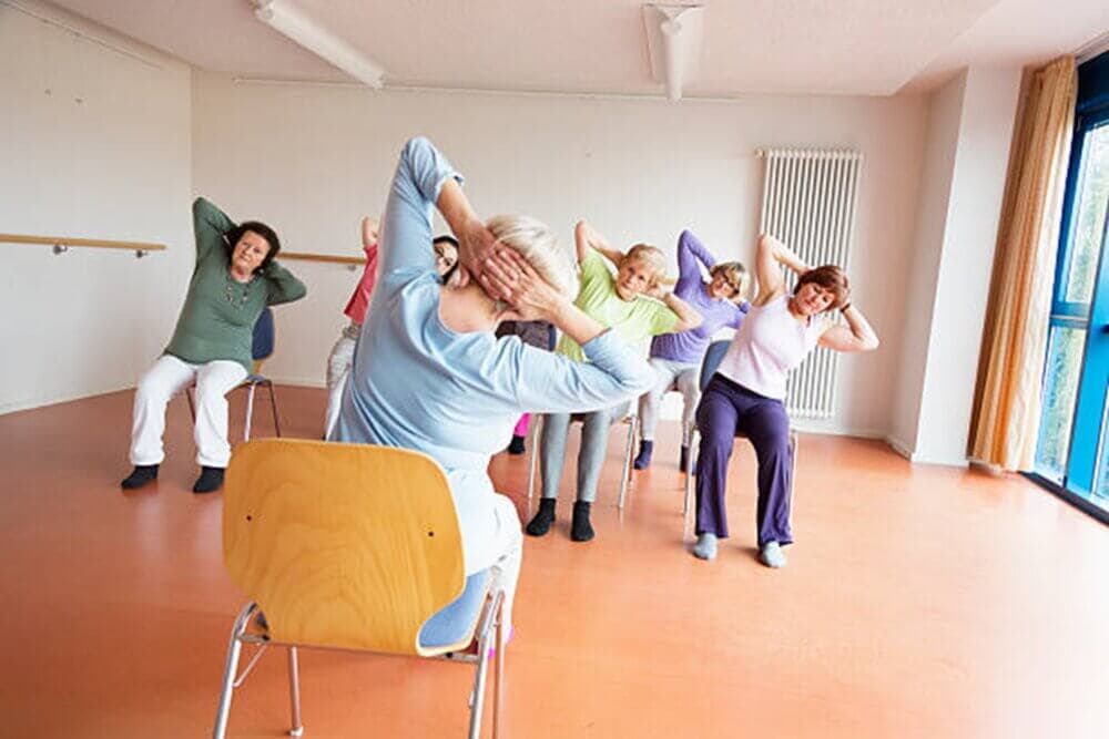 A group of seniors seated on chairs doing stretching exercises in a bright room with large windows. - Home Instead