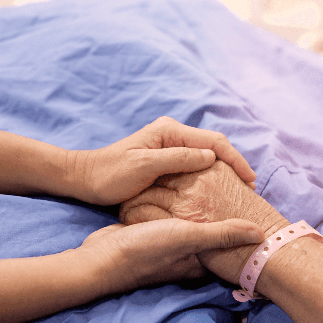 A person holding the hand of an elderly patient lying in a hospital bed, wearing a pink wristband. - Home Instead