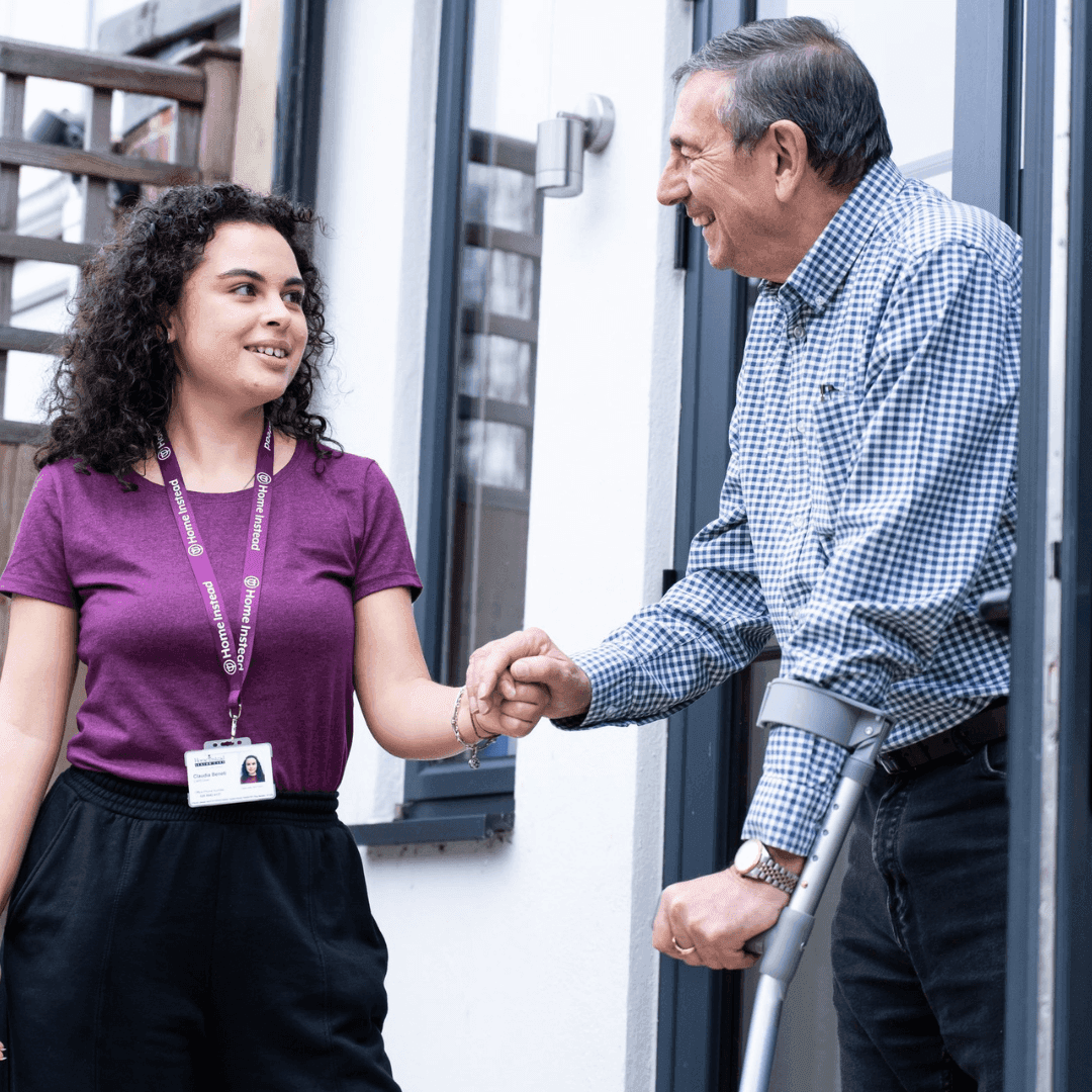 Young woman with a name badge helps an older man with crutches at an entrance, both smiling and shaking hands. - Home Instead