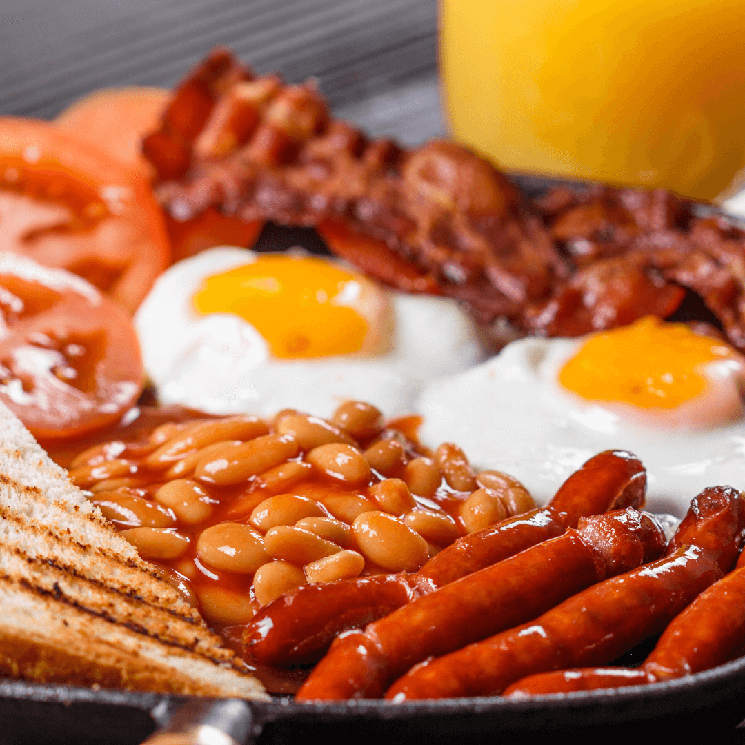 A plate of English breakfast with fried eggs, bacon, sausages, baked beans, tomatoes, toast, and a glass of orange juice. - Home Instead