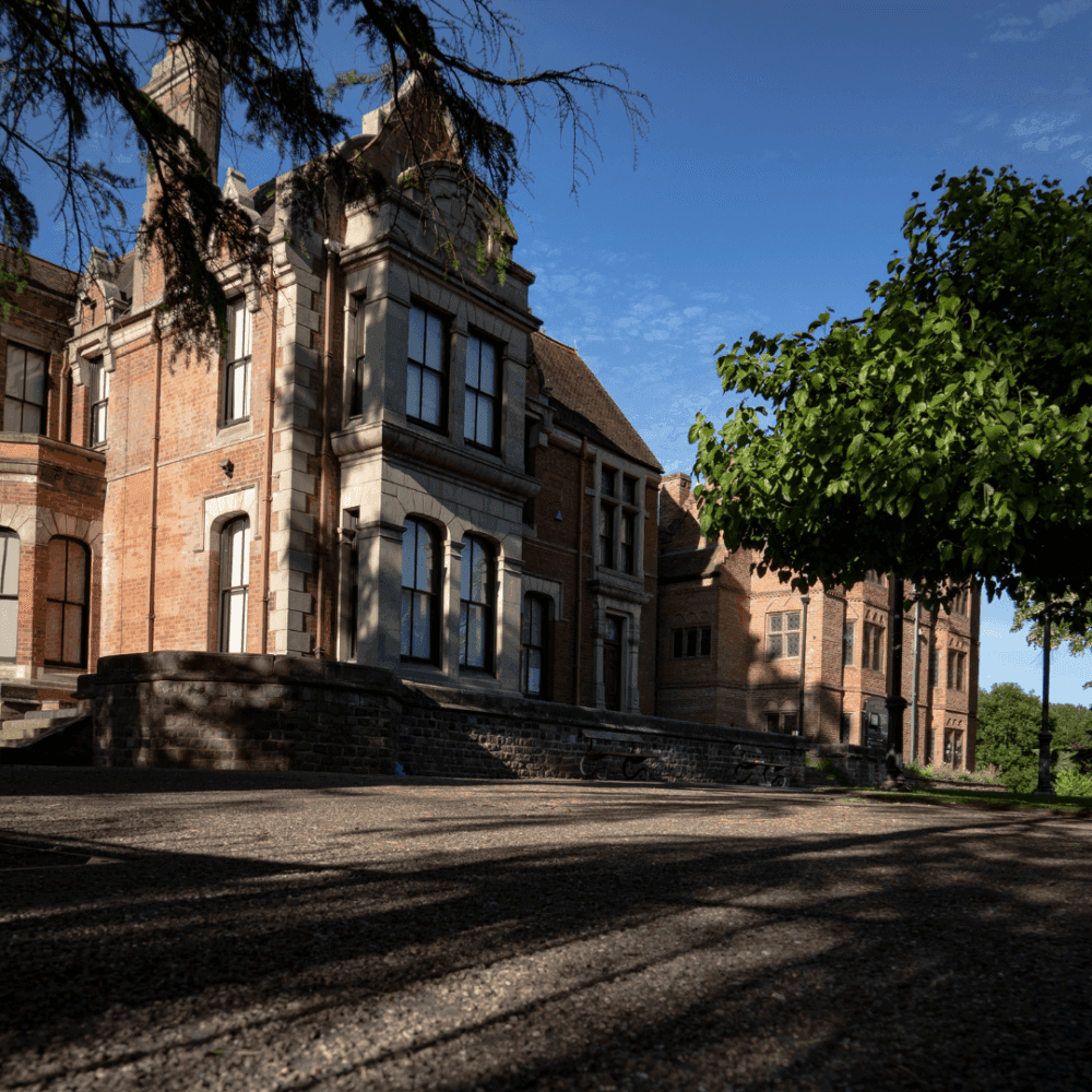 A historic brick building with large windows, framed by a tree branch on the left and lush greenery on the right. - Home Instead