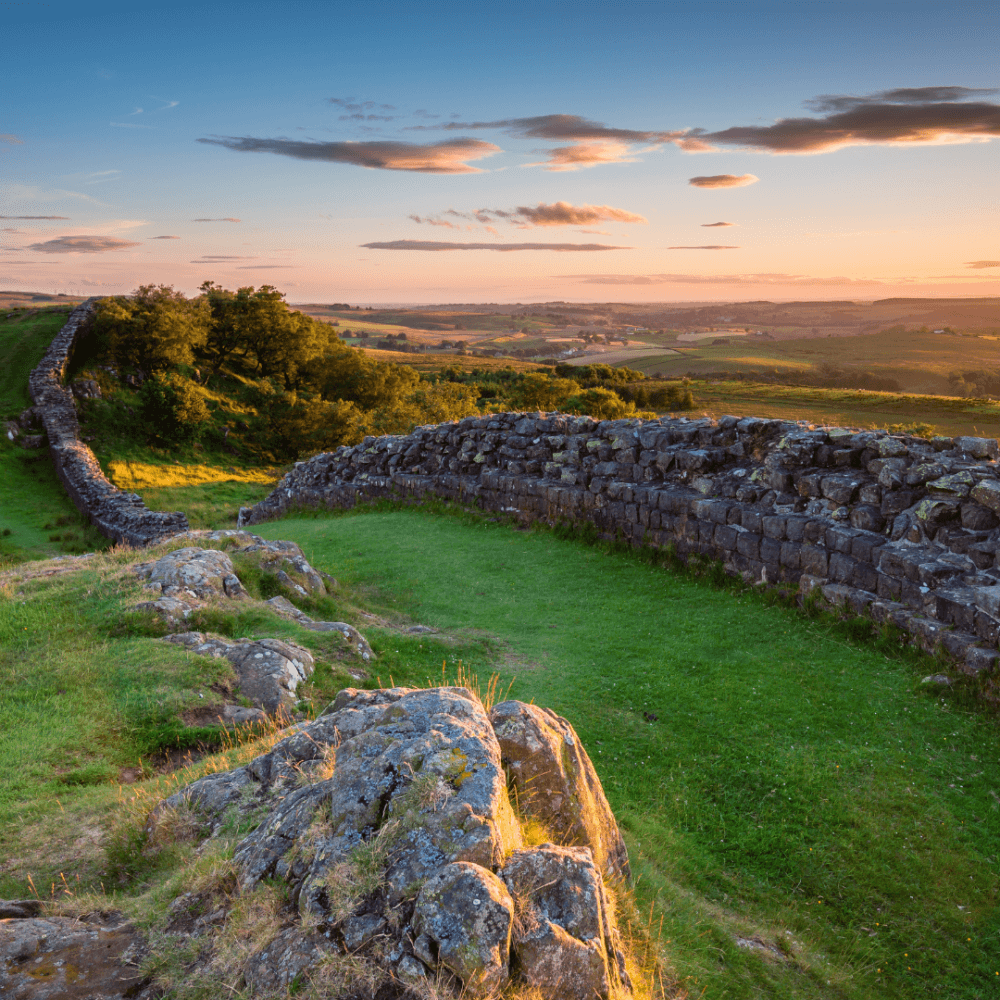 Hadrian's Wall winding through a green landscape at sunset, with a clear blue and orange sky overhead. - Home Instead