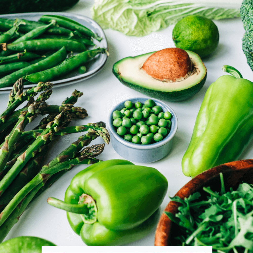Assorted green vegetables including bell peppers, avocado, peas, asparagus, lettuce, and broccoli on a white surface. - Home Instead
