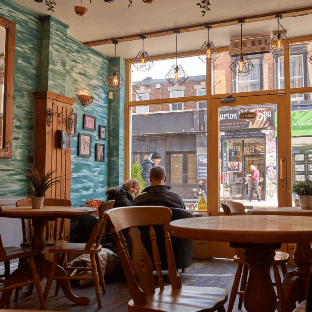 Cozy café with wooden furniture, teal walls, and hanging lights. People sit by the window overlooking a street. - Home Instead