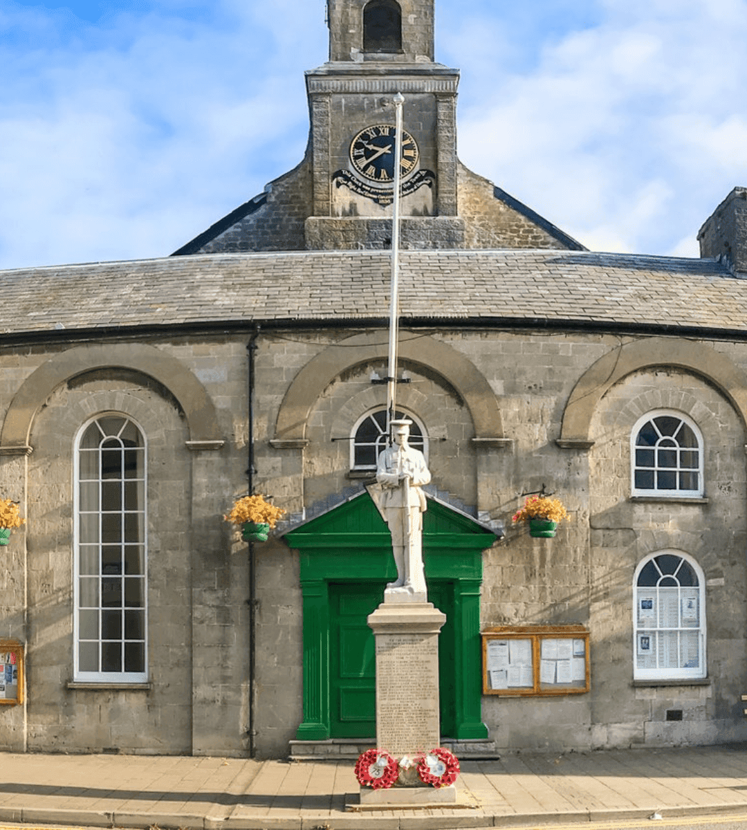 A historical building with arched windows, a clock tower, and a war memorial statue in front. - Home Instead
