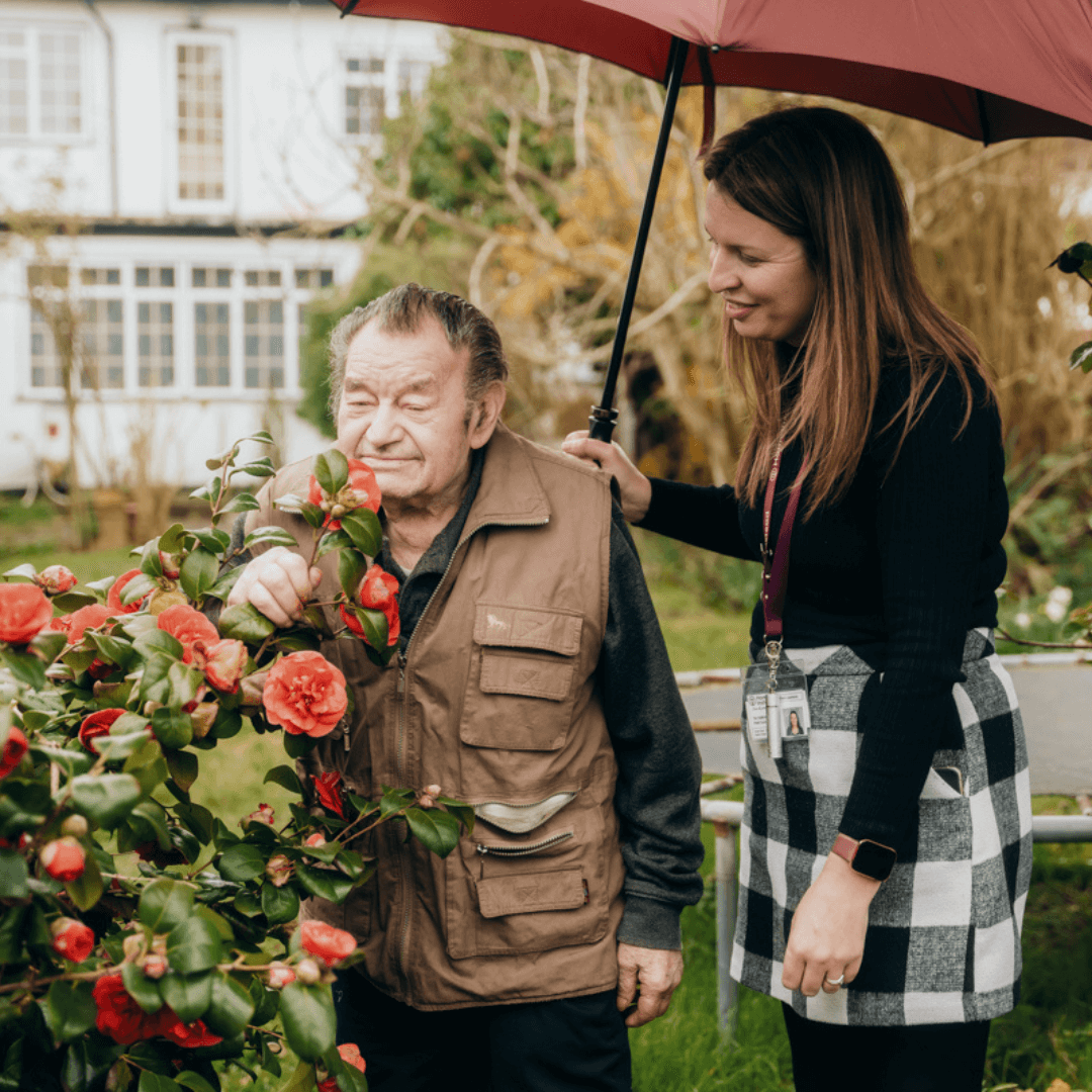 A man inspects flowers while a woman holds a red umbrella over him in a garden. - Home Instead