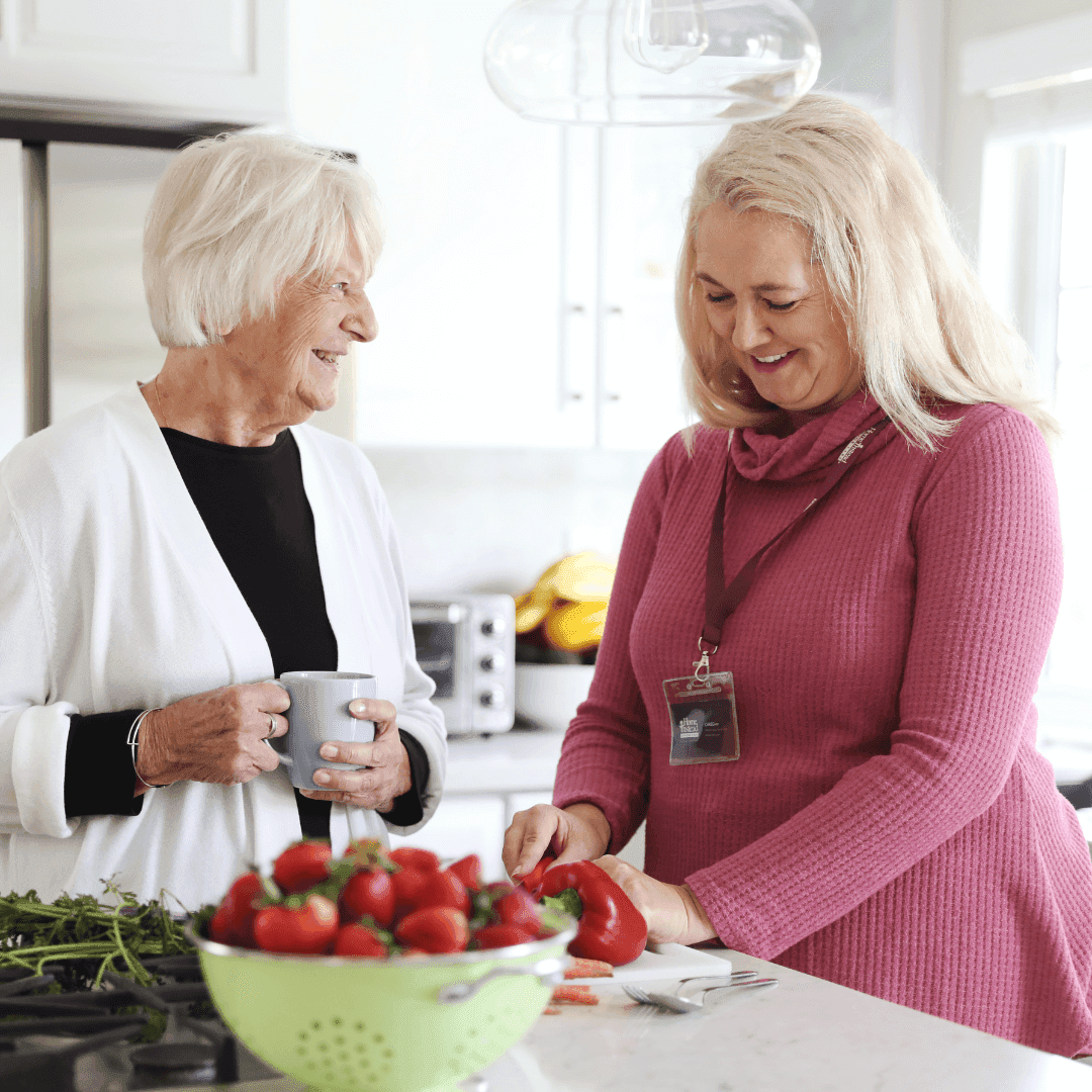 Two women in a kitchen, one elderly holding a mug, and the other younger chopping vegetables with a badge around her neck. - Home Instead